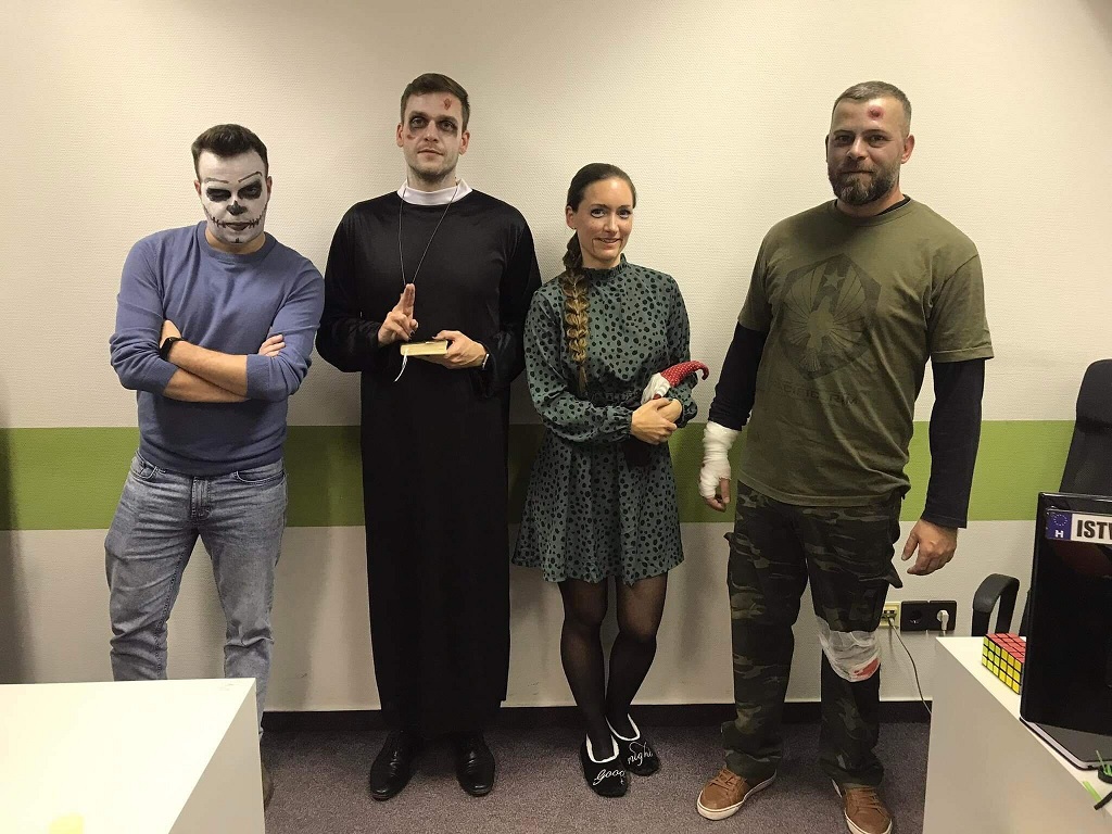 Négy ember egy irodai környezetben pózolnak, Halloween-i jelmezt viselnek.