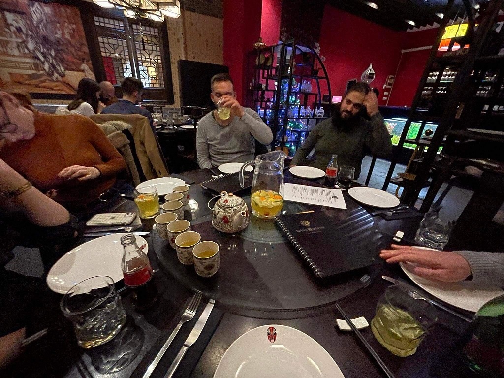 Vacsoraasztal terítéke teáscsészékkel, tányérokkal és étlapokkal egy félhomályos étteremben. Az asztal körül emberek ülnek.