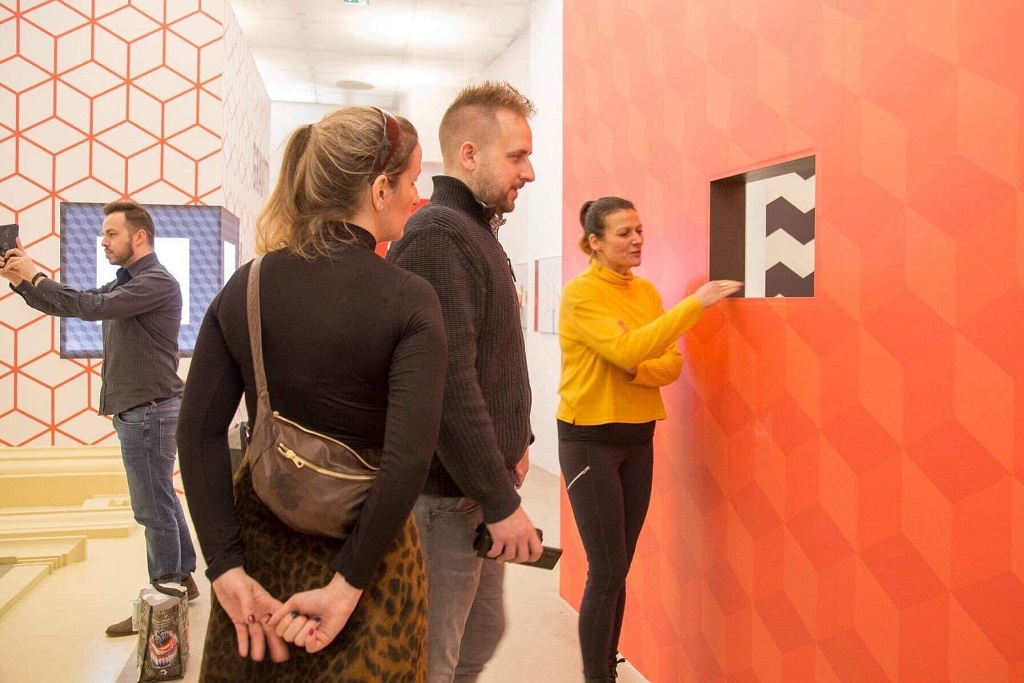 Emberek műalkotásokat nézegetnek egy galériában, amelynek falai geometrikus mintázatúak.